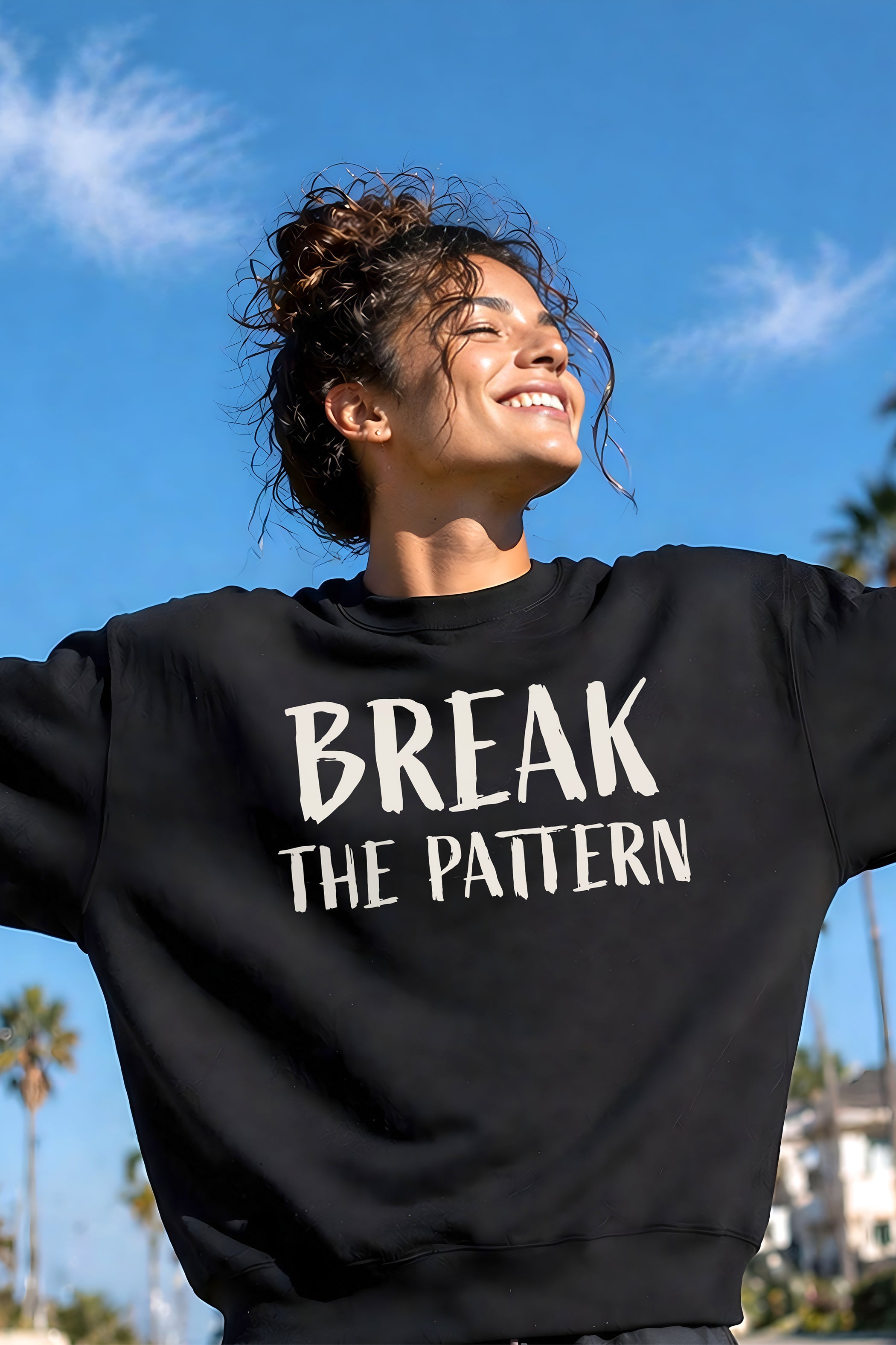 Model standing outside in the sun smiling wearing a black unisex crewneck sweatshirt featuring a bold off white text graphic Break The Pattern, created by content creators Kayla and Samantha.