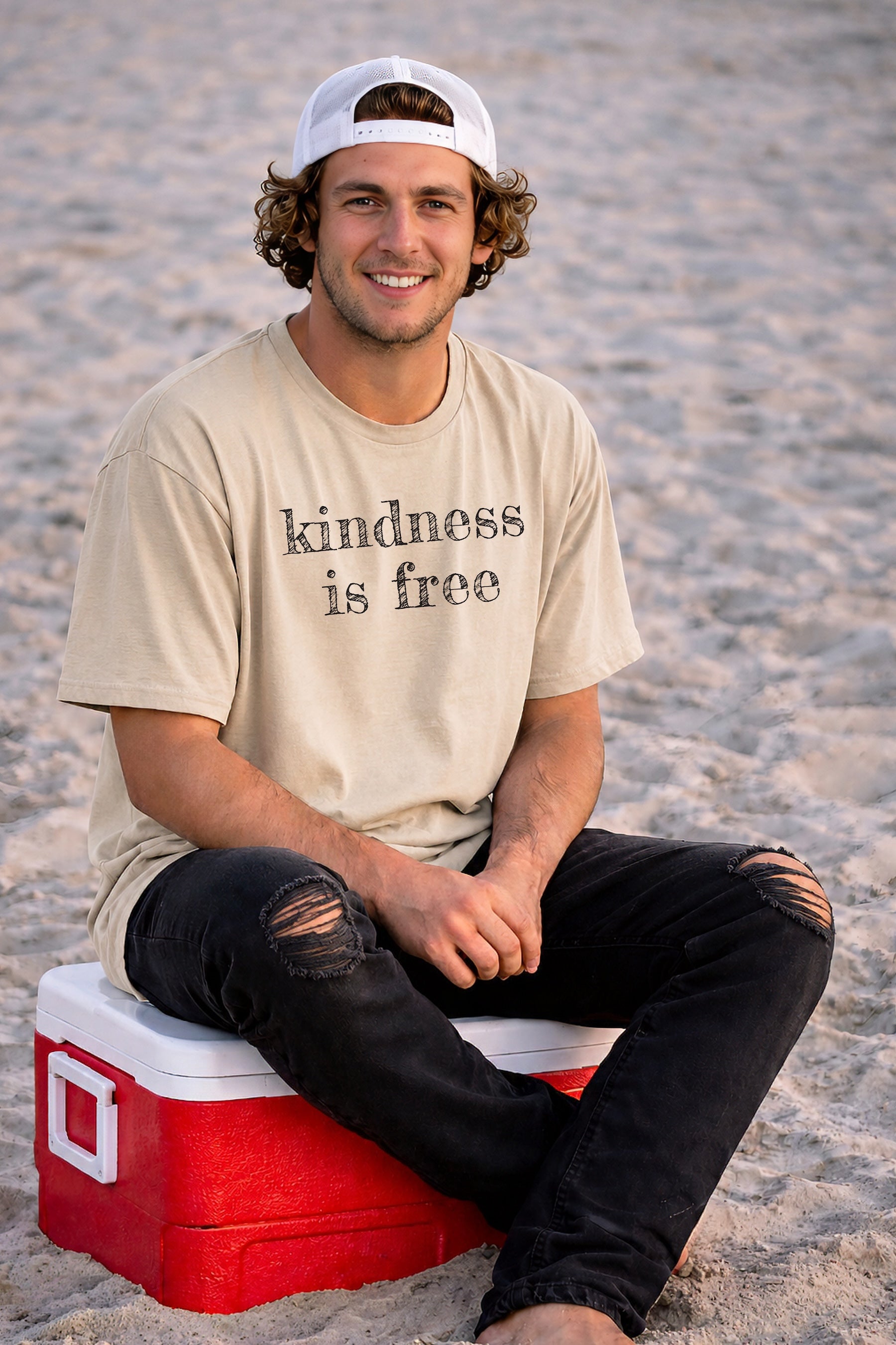 Model sitting on a beach cooler wearing a light blue unisex t-shirt featuring a typewriter text graphic Kindness Is Free, created by content creators Kayla and Samantha.