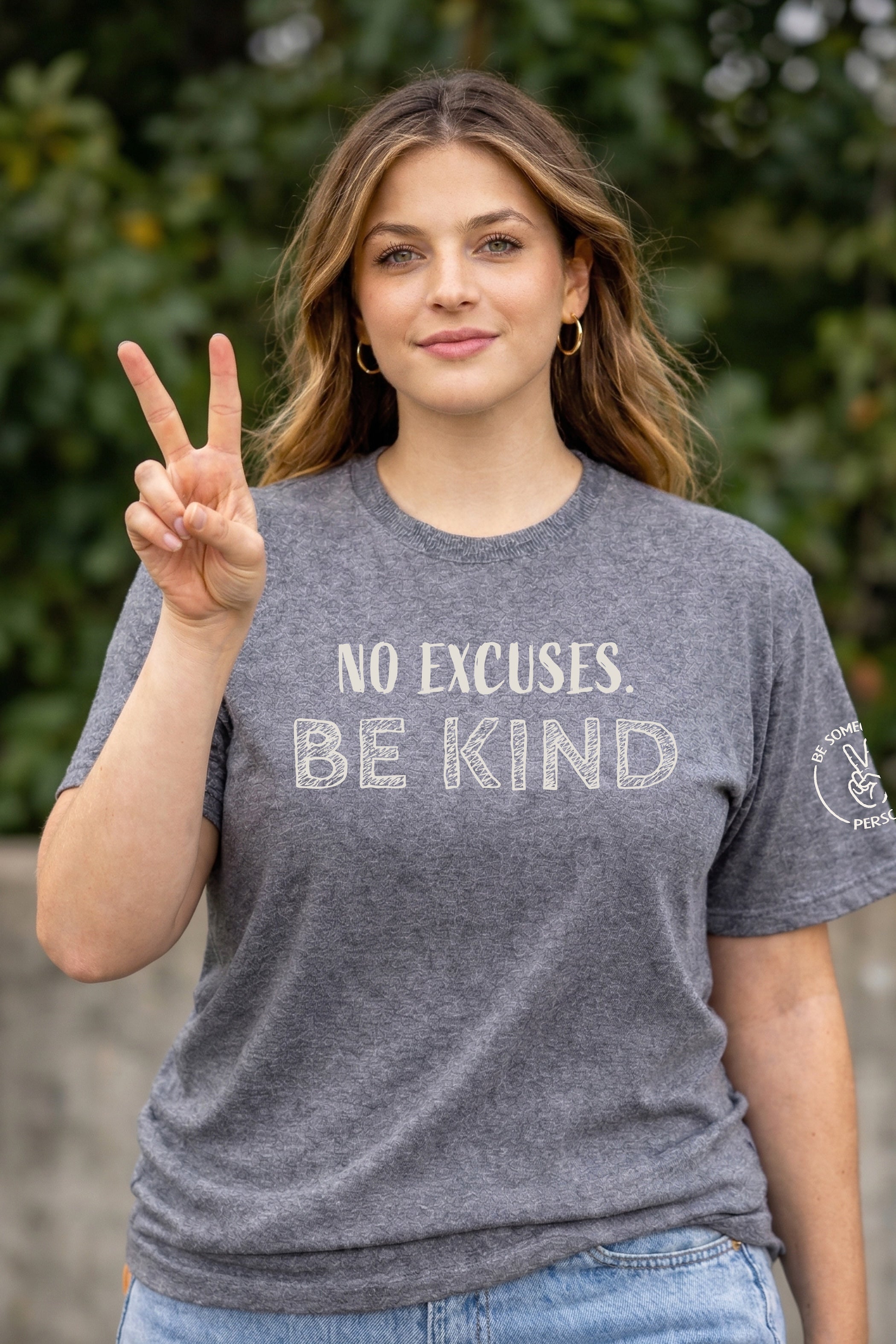 Model wearing a dark gray unisex t-shirt featuring the text graphic No Excuses Be Kind while holding up a peace sign, created by content creators Kayla and Samantha.