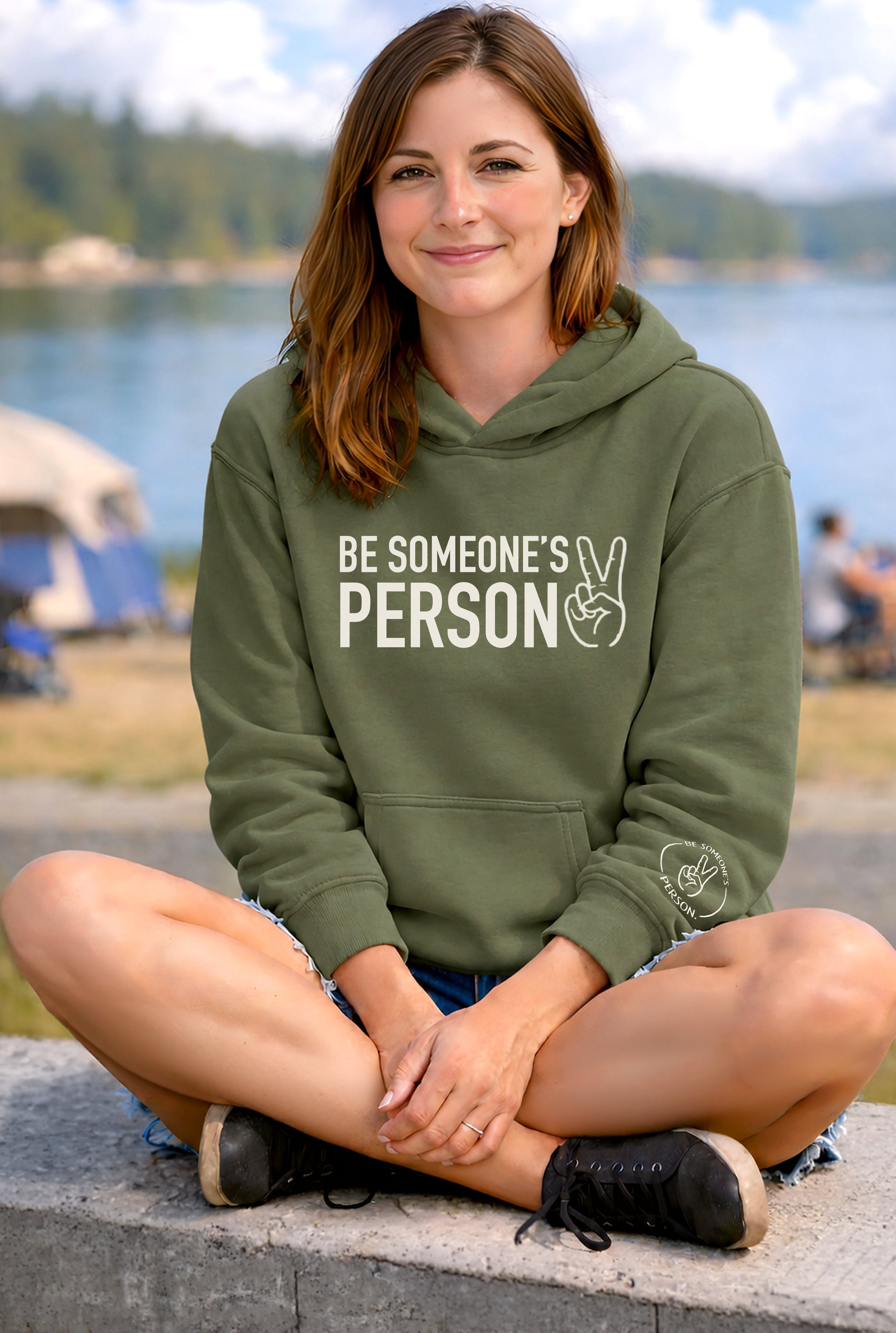 model sitting by a lake smiling wearing a light pink hoodie with the be someones person logo front design, created by kayla and samantha