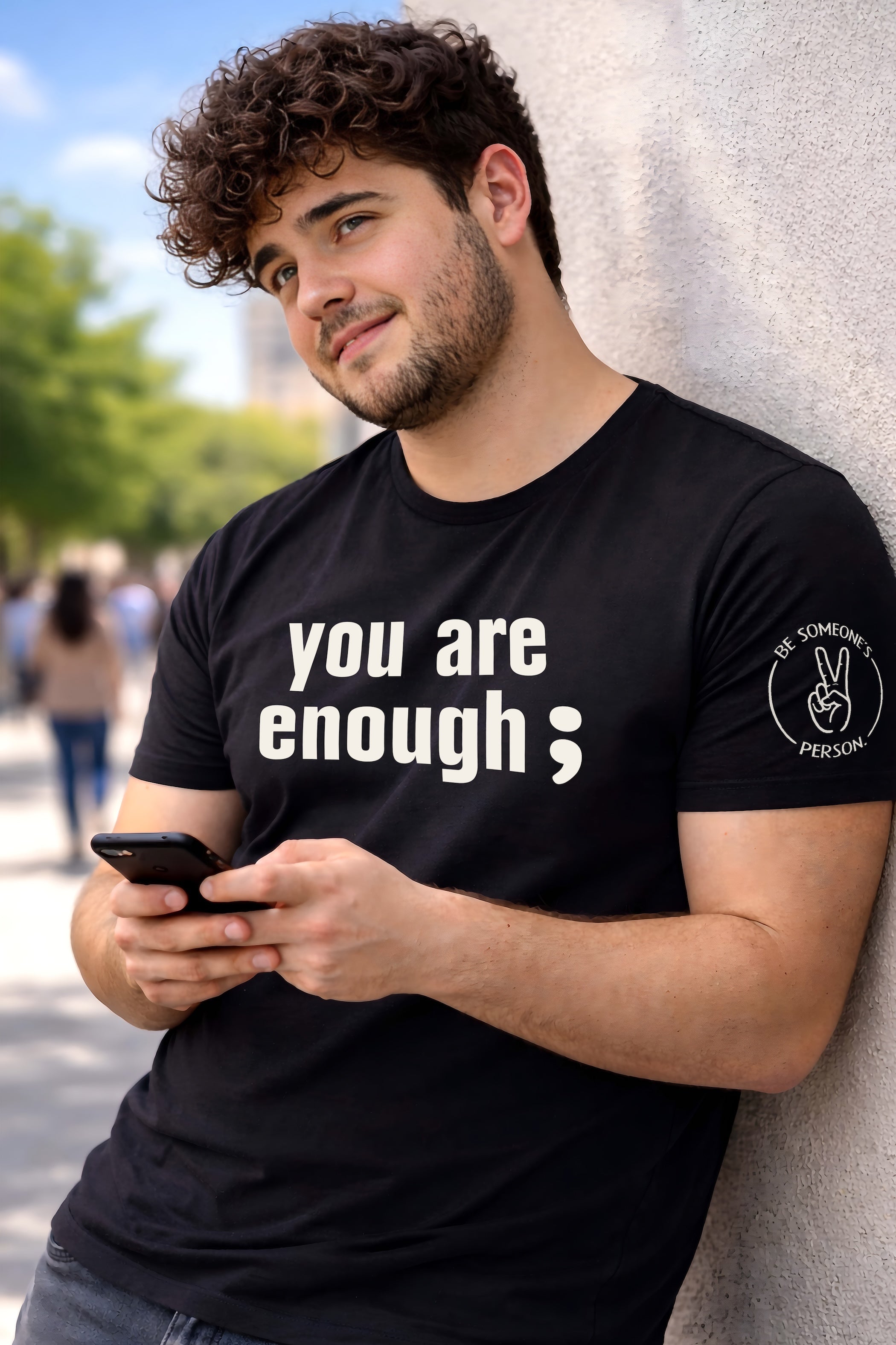 Model leaning against a wall wearing a black unisex t-shirt featuring the text graphic You Are Enough with a semicolon, created by content creators Kayla and Samantha.
