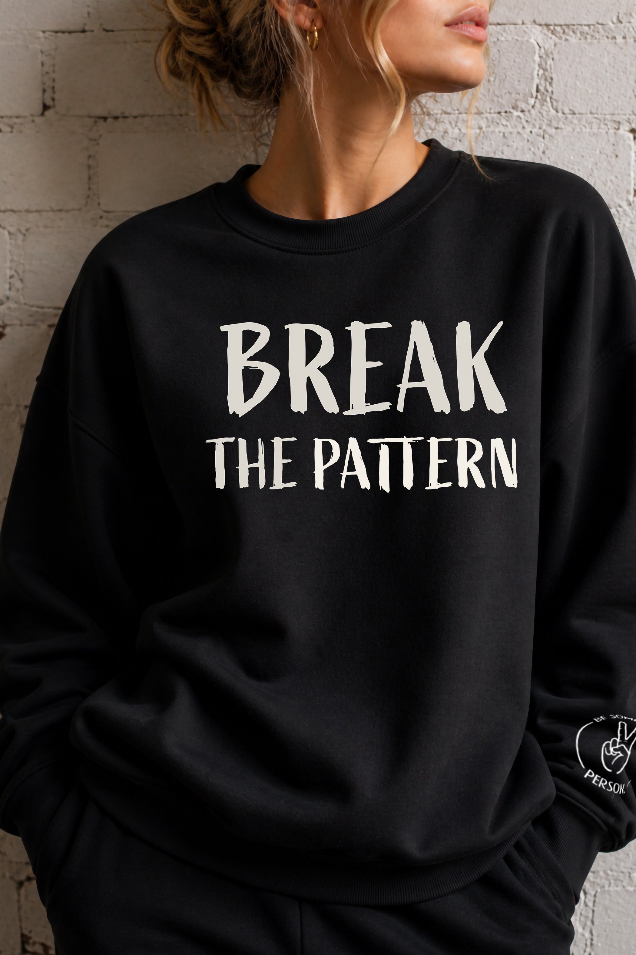 model standing up against a wall relaxed and wearing a black crewneck with the off white text graphic that reads Break The Pattern, created by kayla and samantha for the be someones person brand
