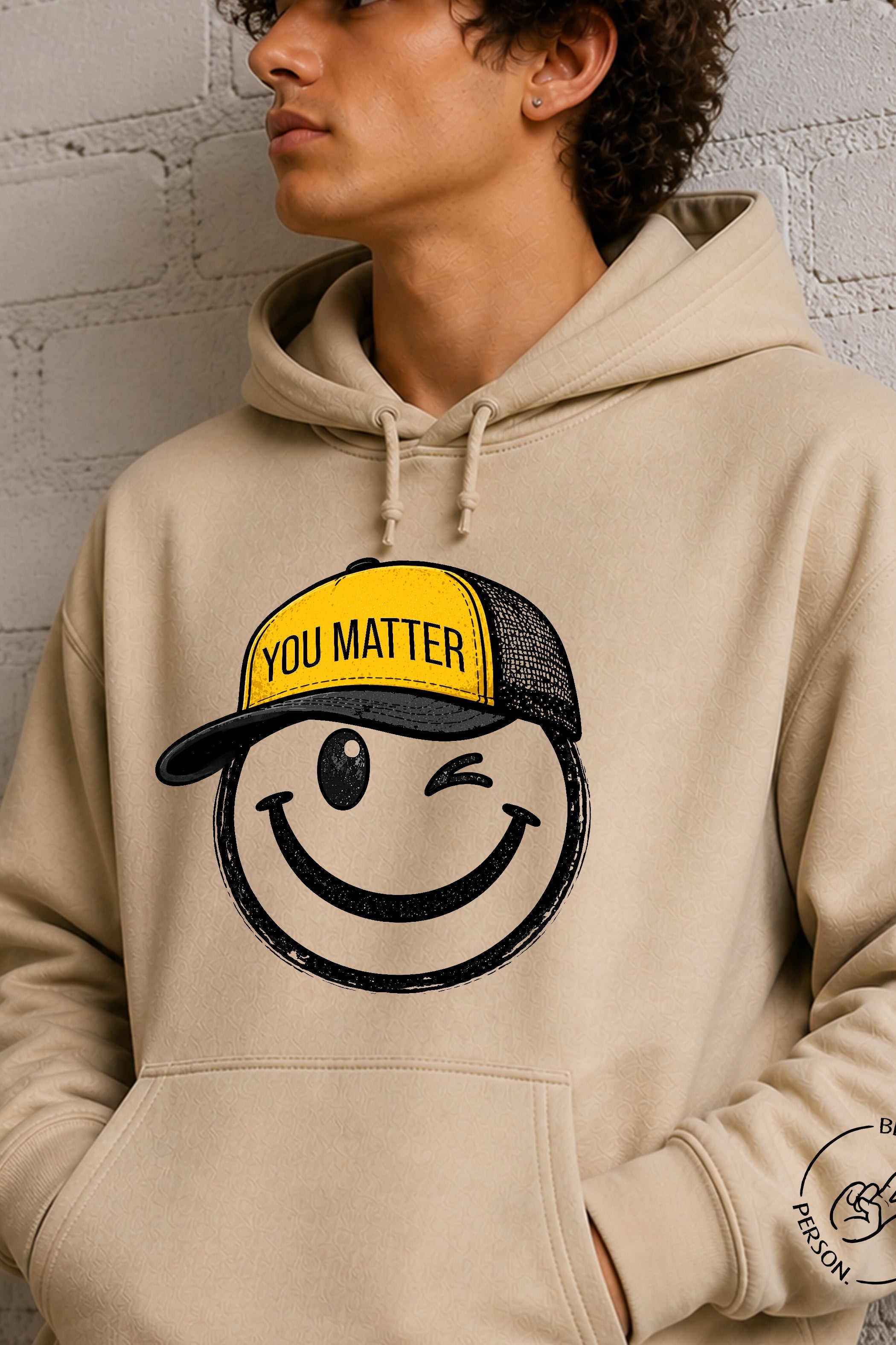 Model wearing a sand unisex hoodie featuring the winking You Matter smiley face logo from Be Someone's Person, created by content creators Kayla and Samantha.